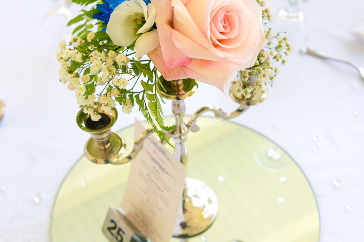 Centre de table floral avec une rose rose, des fleurs blanches et bleues, posé sur un miroir rond avec un numéro 25.