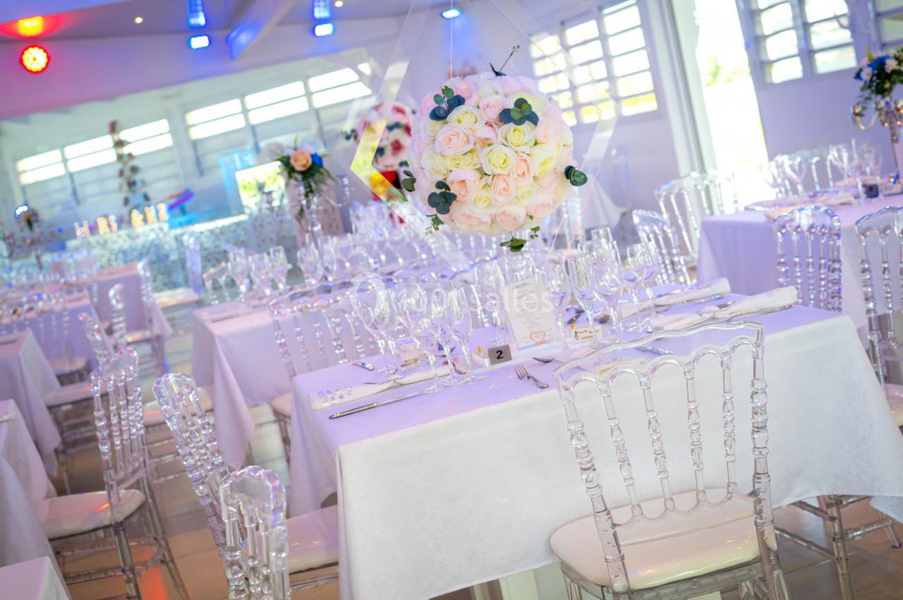 Salle de réception lumineuse avec tables dressées, nappes blanches, chaises transparentes et centre de table floral.