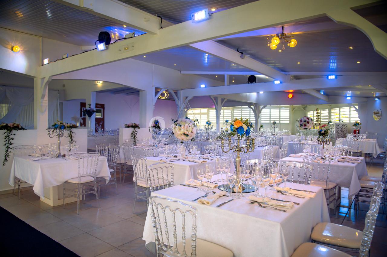Salle de réception lumineuse avec tables décorées de nappes blanches, chandeliers et arrangements floraux.