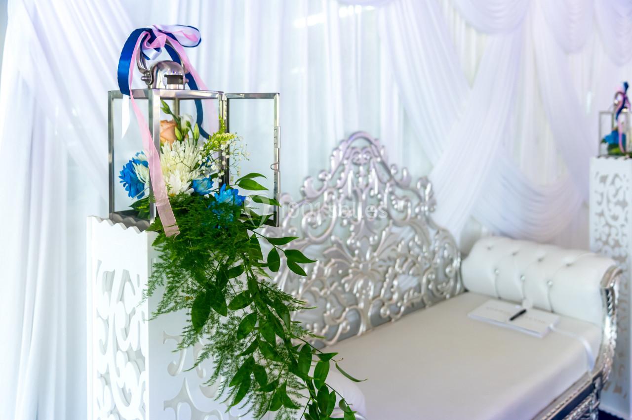 Décoration florale avec des rubans et des feuillages sur un support blanc, devant un canapé argenté capitonné.