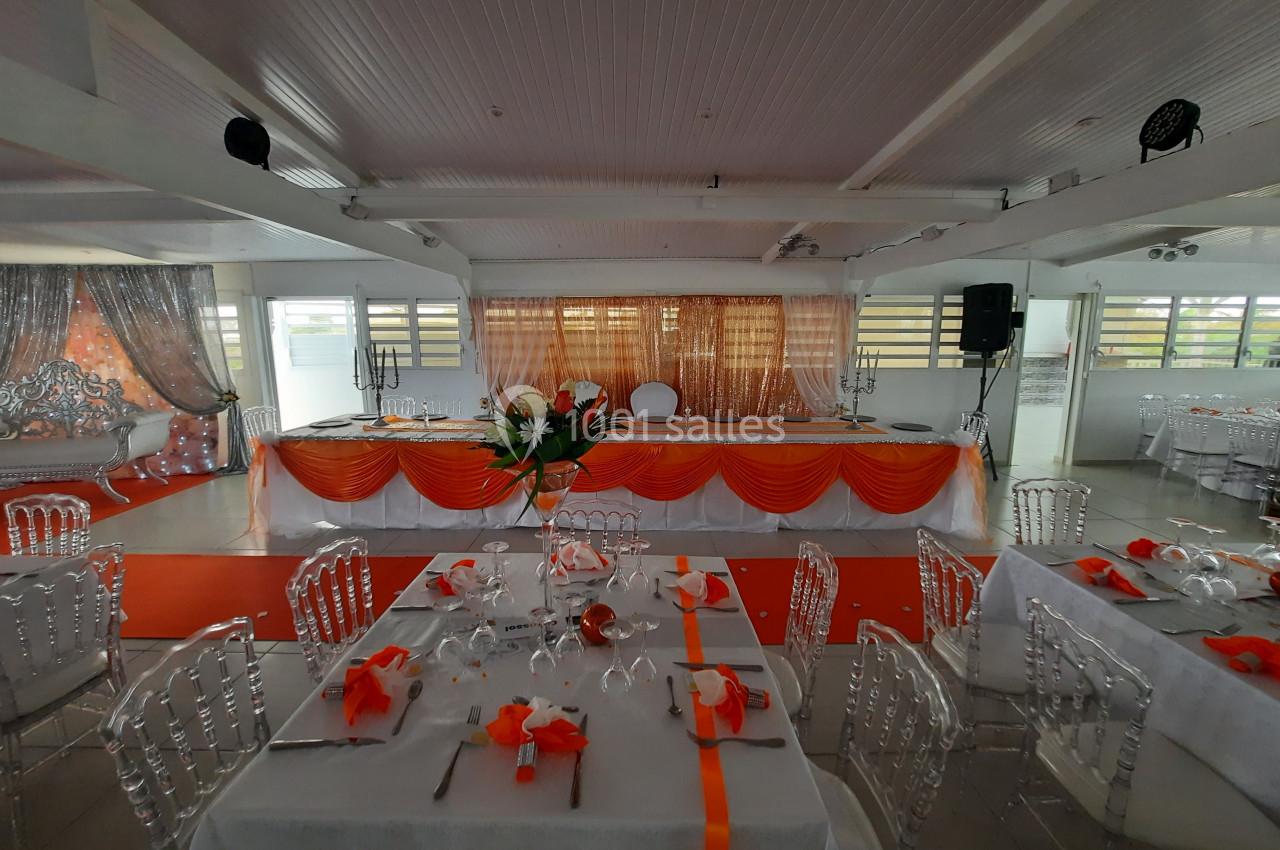 Salle de réception décorée avec des nappes blanches et des accents orange, tables dressées et estrade ornée en arrière-plan.