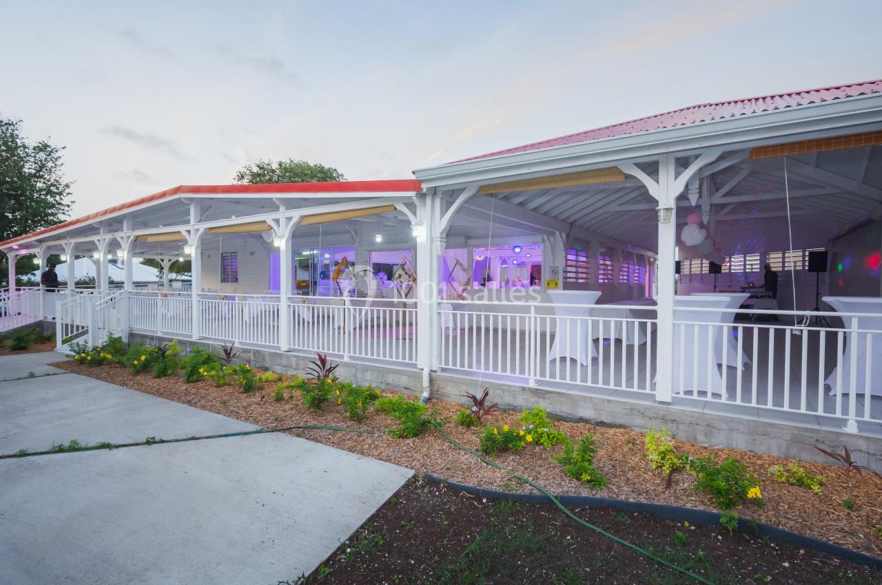Salle de réception ouverte avec toit rouge, tables hautes blanches et éclairage intérieur violet, entourée de végétation.