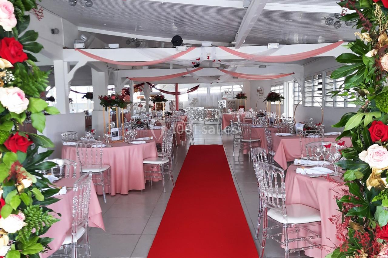Salle de réception décorée avec des tables roses, des chaises transparentes et un tapis rouge central.