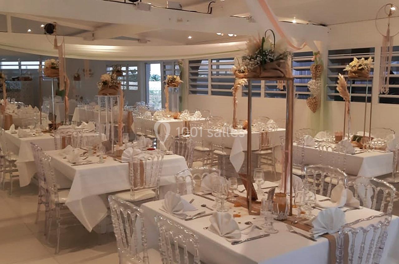 Salle de réception décorée avec des tables dressées, nappes blanches, chaises transparentes et ornements floraux suspendus.