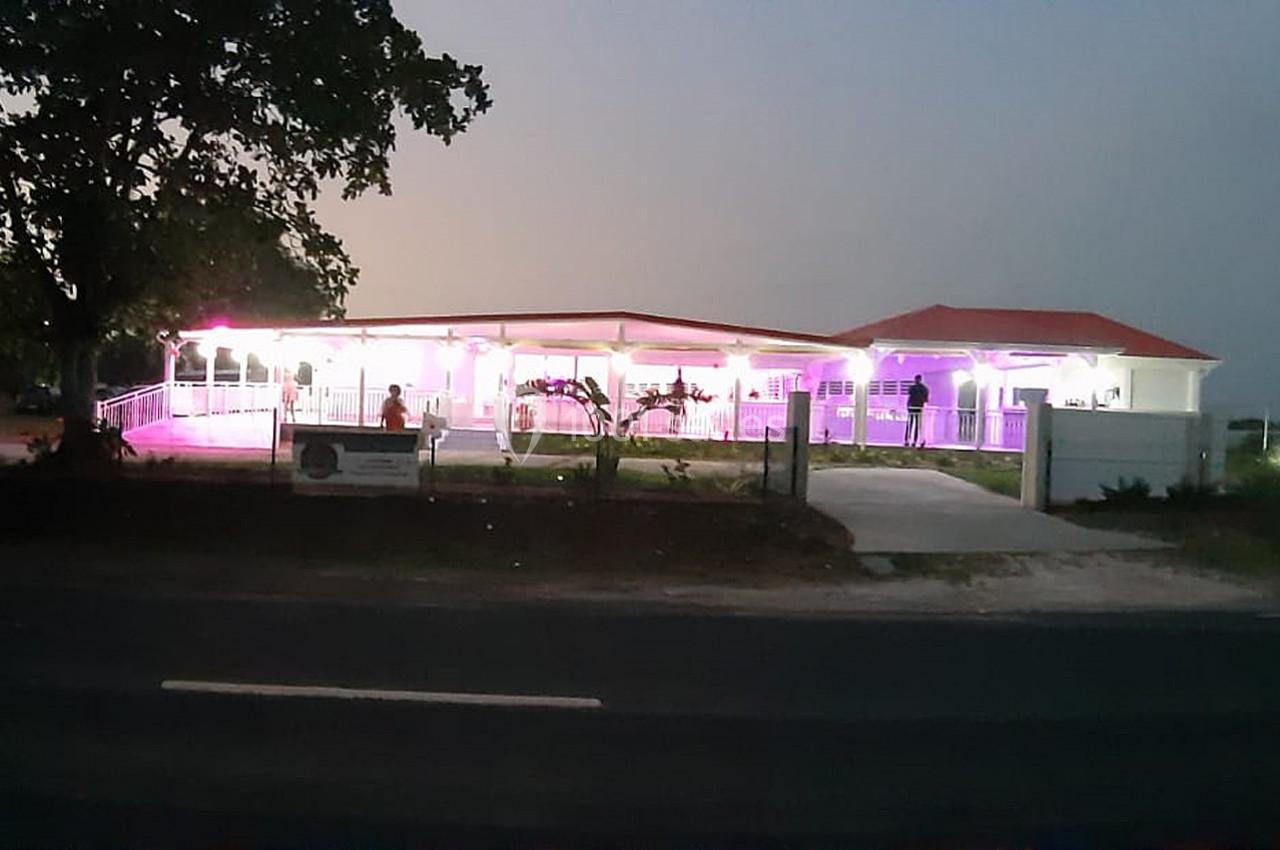 Bâtiment éclairé en rose et blanc à la tombée de la nuit, entouré d'arbres et situé près d'une route.