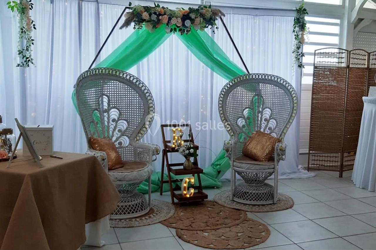 Deux fauteuils en rotin avec coussins dorés, décorés de fleurs et d'un drapé vert, dans un espace événementiel.