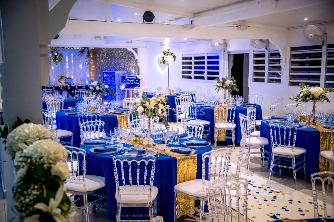 Salle de réception décorée avec des tables rondes, nappes bleues, chaises transparentes et arrangements floraux élégants.