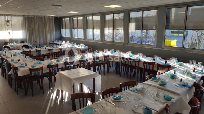 Salle de réception lumineuse avec tables dressées, nappes blanches et serviettes bleues, prête pour un événement.