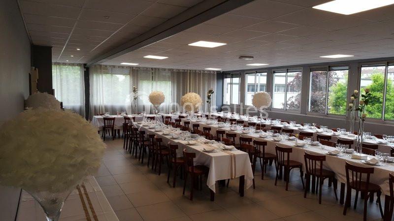 Salle de réception lumineuse avec tables alignées, nappes blanches et décorations florales blanches.