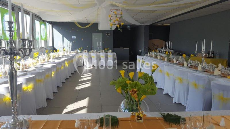 Salle décorée pour un événement avec tables alignées, nappes blanches, centres de table floraux et guirlandes au plafond.