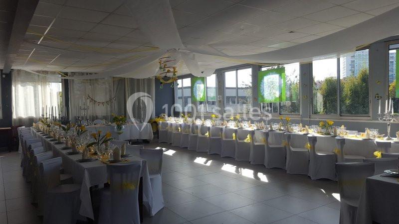 Salle de réception décorée avec des tables alignées, nappes blanches et accents jaunes, éclairée par la lumière naturelle.