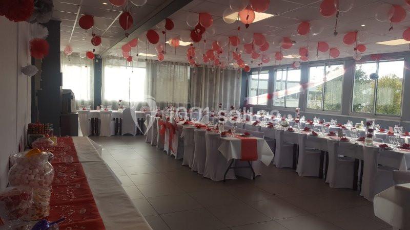 Salle de réception décorée avec des ballons rouges et blancs, tables dressées avec nappes et chemins de table assortis.
