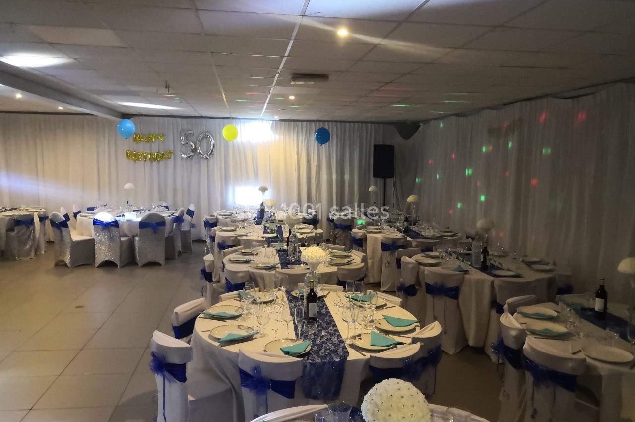 Salle décorée pour une fête avec tables dressées, nappes blanches, rubans bleus et ballons suspendus.