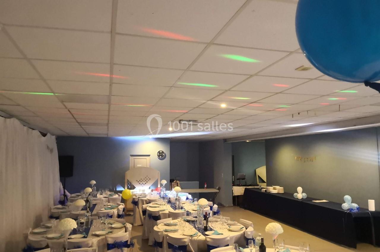 Salle de réception décorée avec des tables dressées, nappes blanches et éléments bleus, éclairage tamisé.