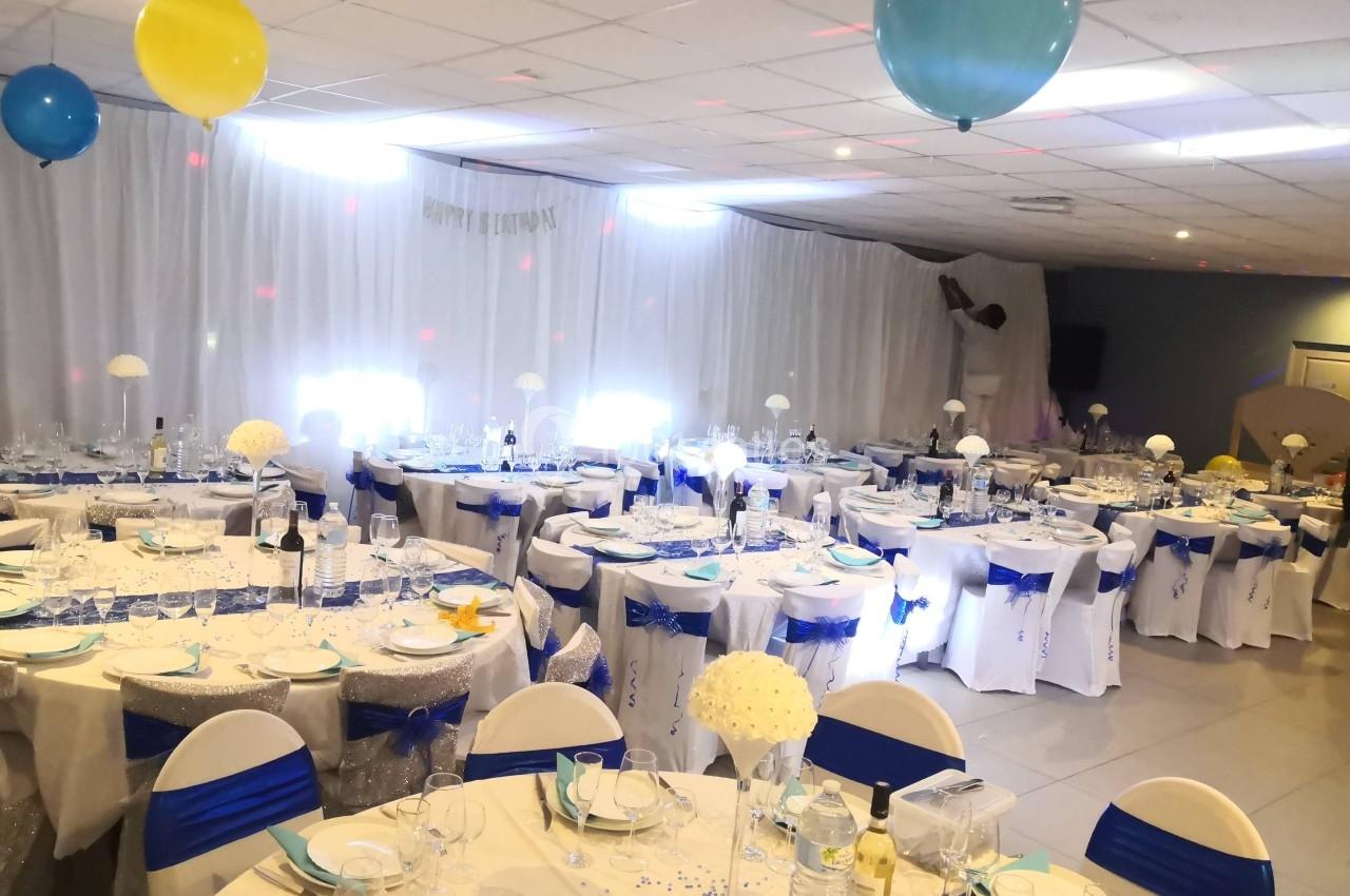 Salle de réception décorée avec des tables rondes, nappes blanches, rubans bleus et ballons suspendus au plafond.