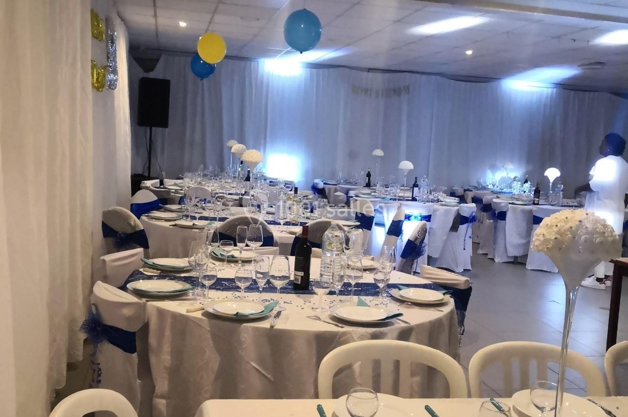 Salle décorée pour un événement avec tables dressées, nappes blanches, chemins de table bleus et ballons suspendus.