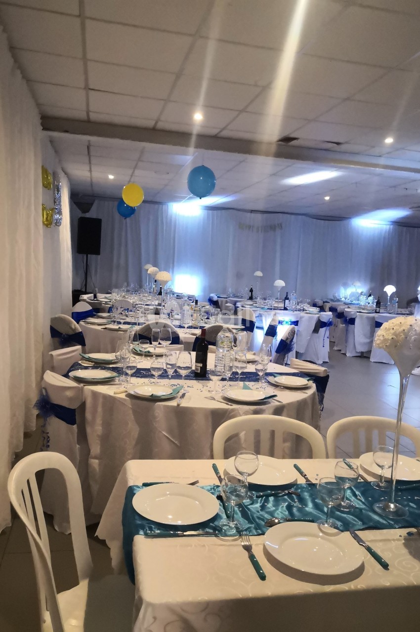 Salle décorée pour un événement avec tables dressées, nappes blanches, chemins de table bleus et ballons suspendus.