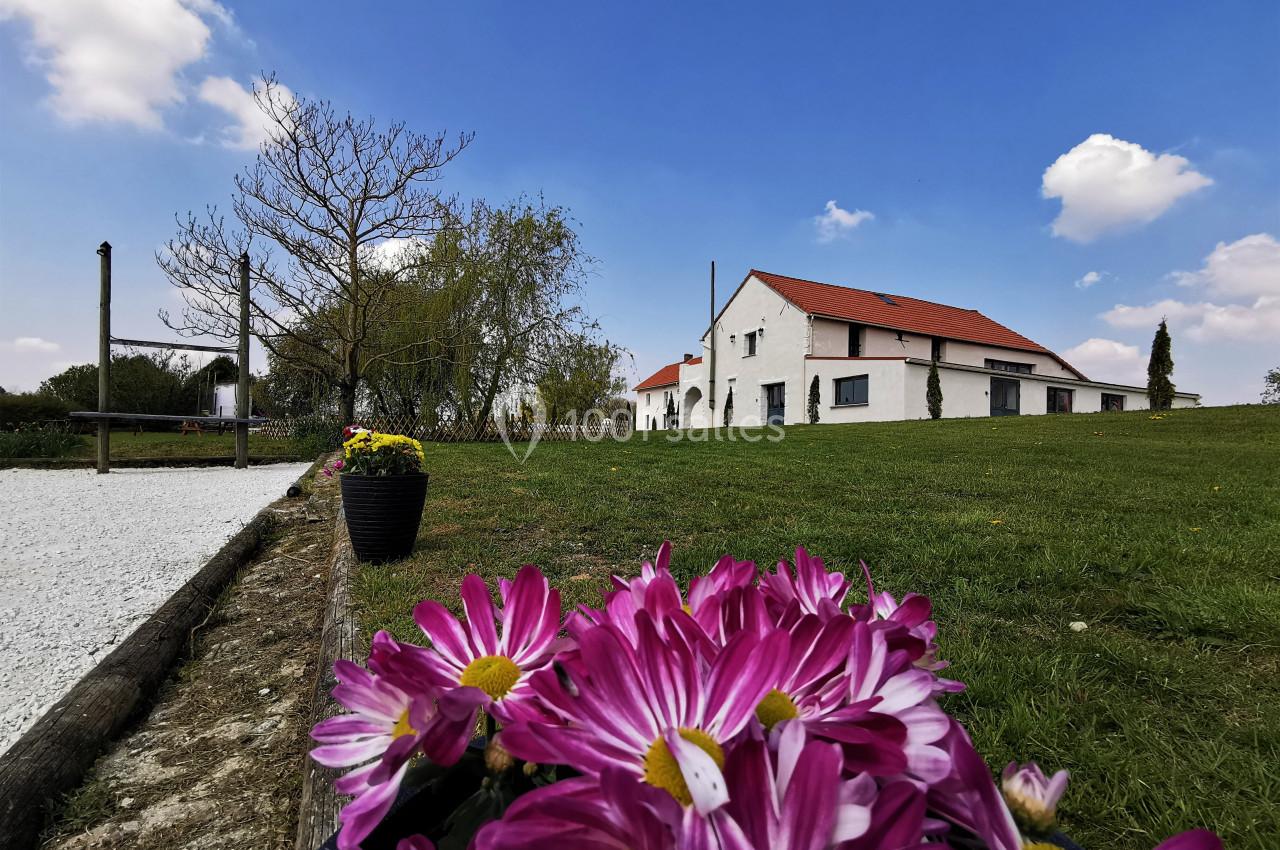 Maison blanche avec toit rouge entourée de pelouse, vue depuis des fleurs roses au premier plan sous un ciel bleu.