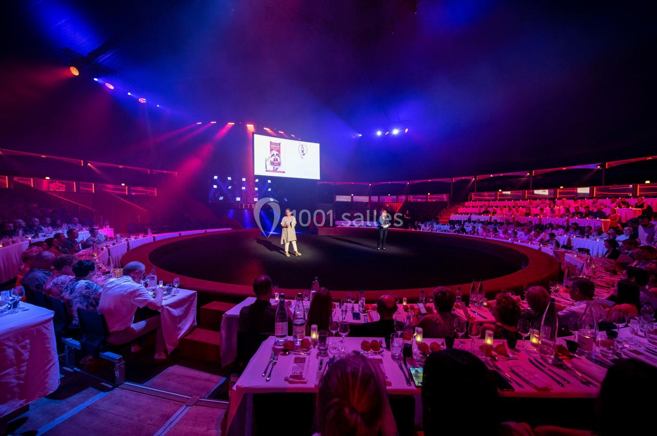 Deux personnes sur une scène circulaire éclairée, entourée de spectateurs assis à des tables dans une ambiance de spectacle.