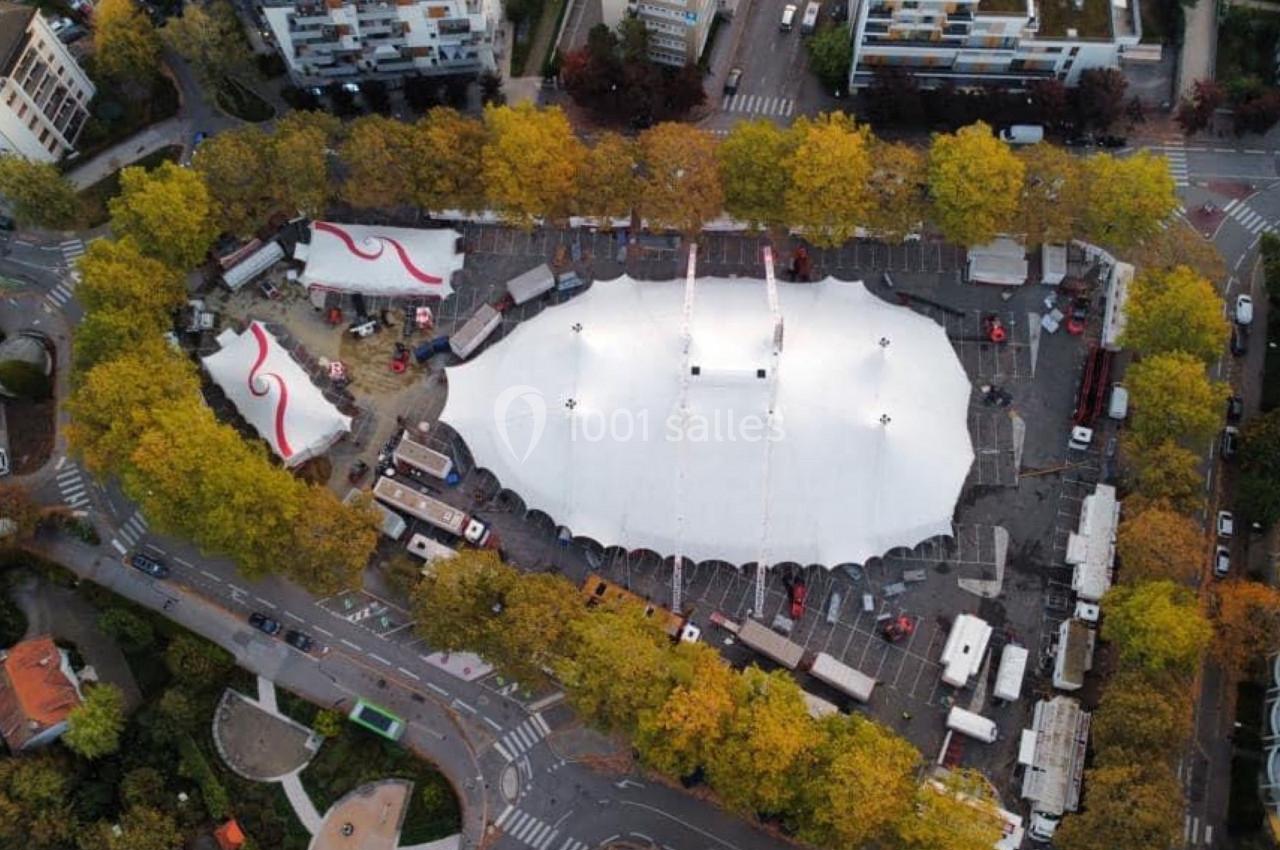 Vue aérienne d'un grand chapiteau blanc entouré d'arbres et de bâtiments dans un espace urbain.