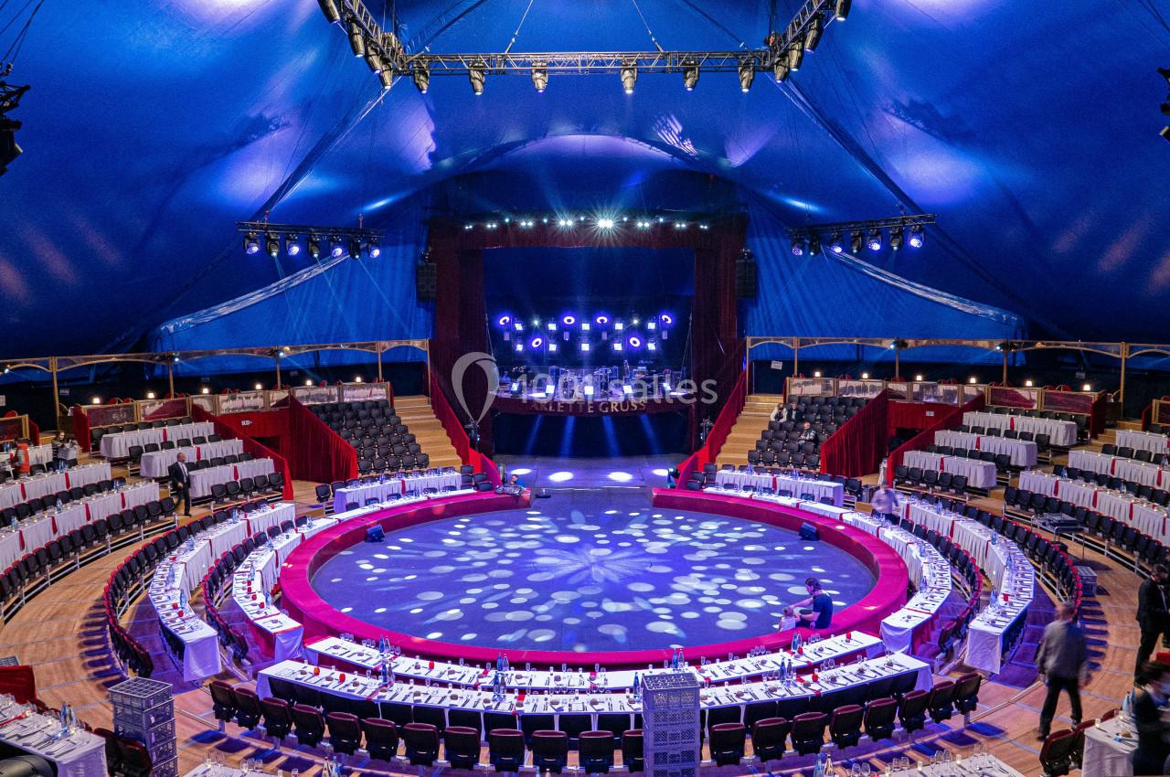 Vue intérieure d'un chapiteau de cirque avec une scène éclairée, des tables dressées et des gradins disposés en cercle.