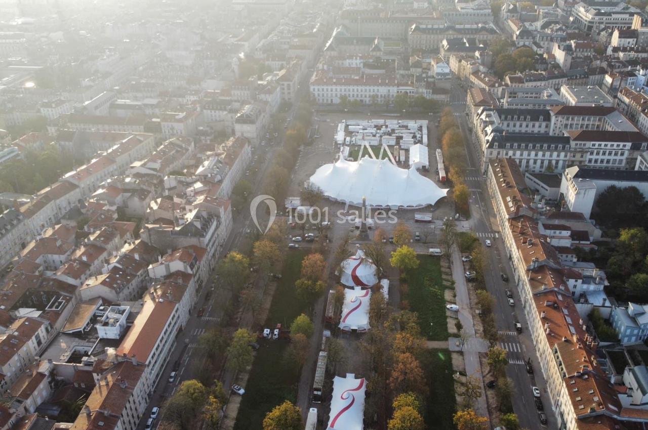 Vue aérienne d'une ville avec des bâtiments, un grand chapiteau blanc et des structures colorées dans un parc.