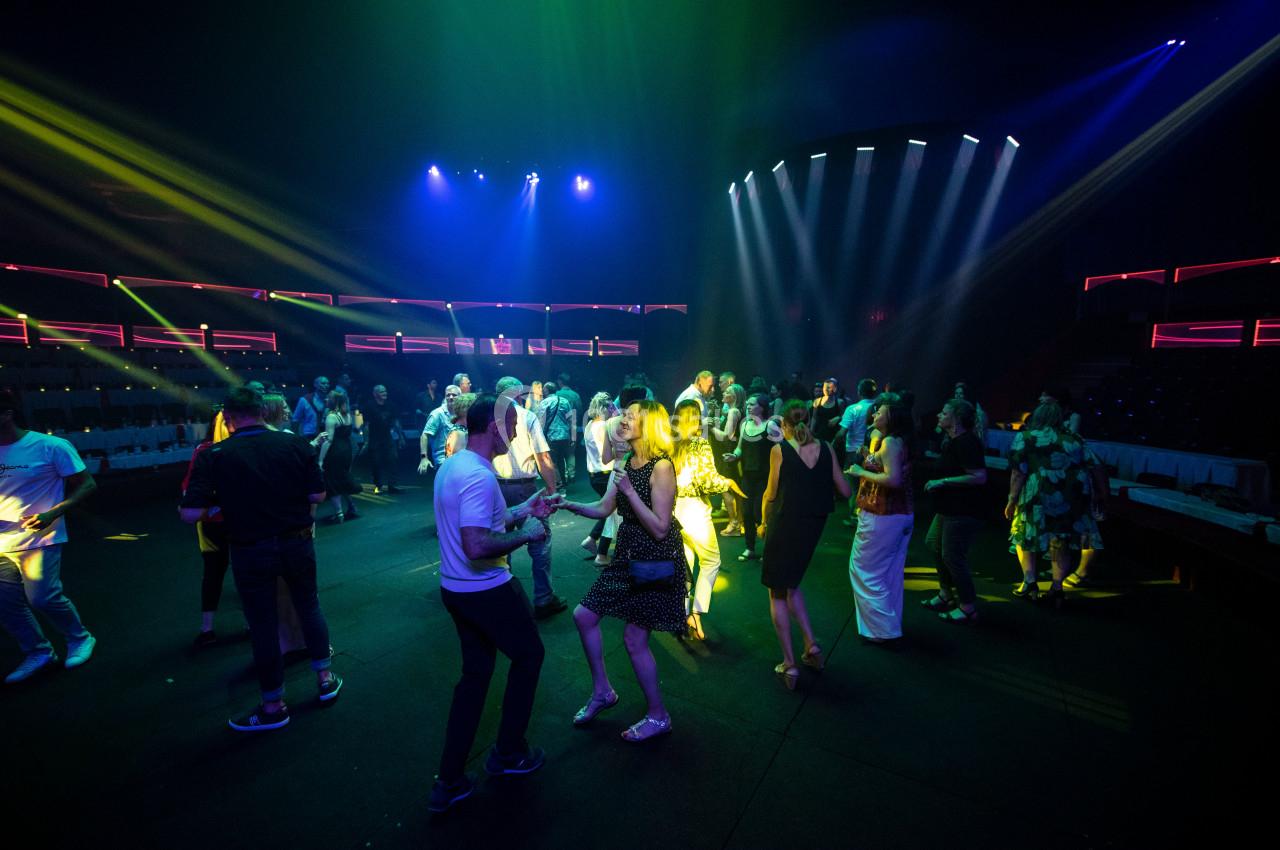 Personnes dansant sous des éclairages colorés dans une salle sombre avec des gradins en arrière-plan.