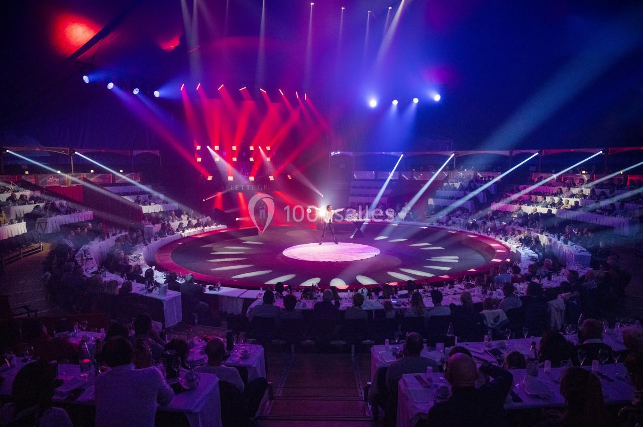 Un artiste se produit sur une scène circulaire éclairée, entourée de spectateurs assis à des tables.