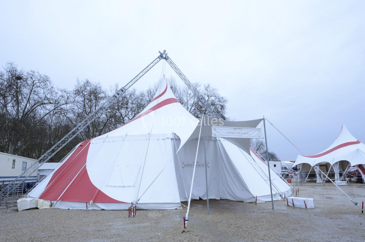 Grand chapiteau blanc et rouge installé sur un terrain gravillonné, entouré d'arbres et de structures similaires.