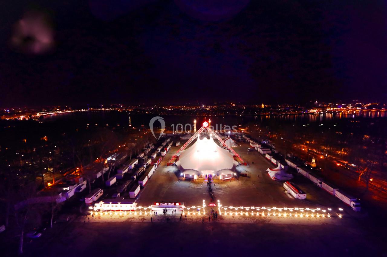 Vue aérienne nocturne d'un grand chapiteau illuminé entouré de stands et de lumières, avec une ville en arrière-plan.