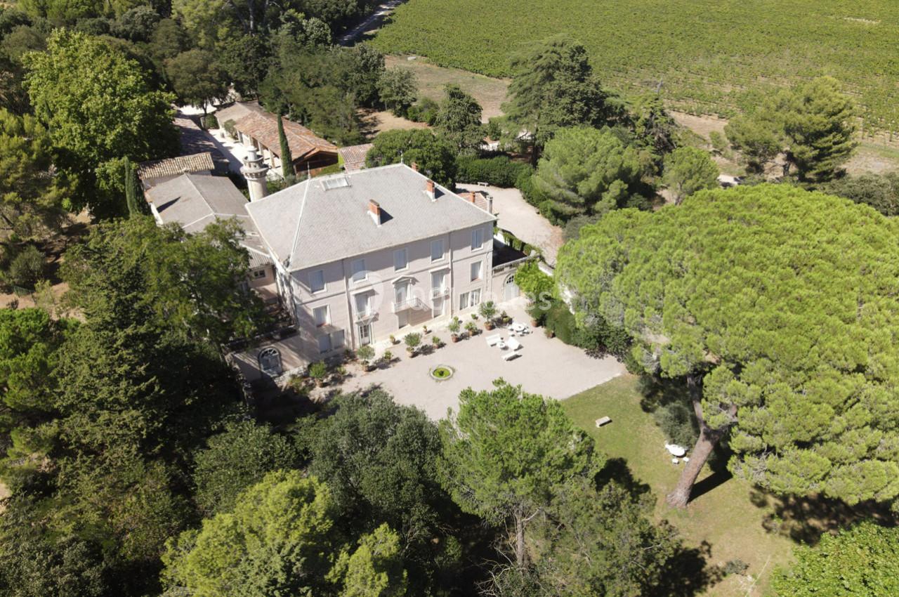 Vue aérienne d'une grande maison entourée d'arbres, avec une cour aménagée et des vignes en arrière-plan.