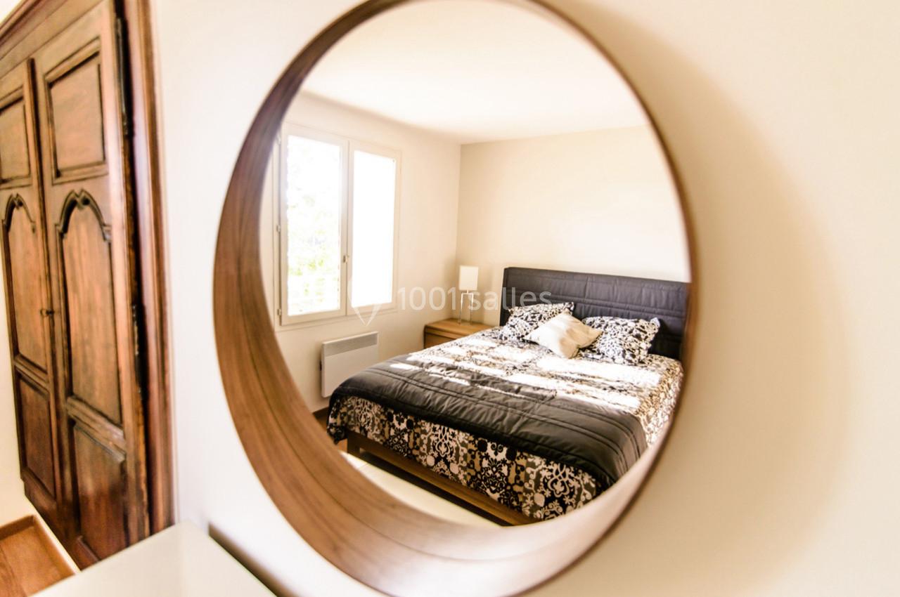 Reflet d'une chambre lumineuse avec un lit double et une armoire en bois, vu à travers un miroir rond.