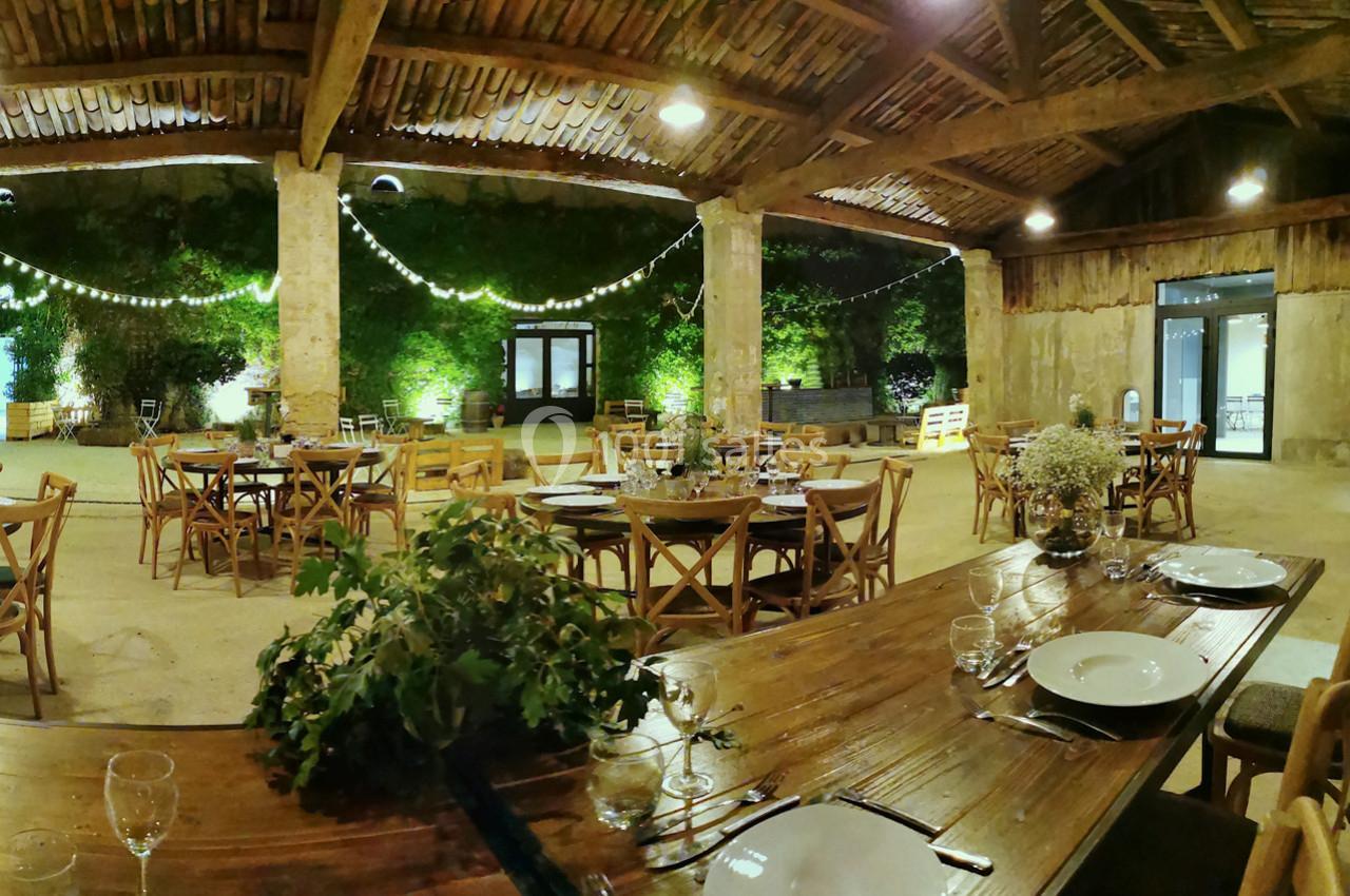 Salle de réception rustique avec tables en bois, vaisselle dressée et guirlandes lumineuses suspendues.