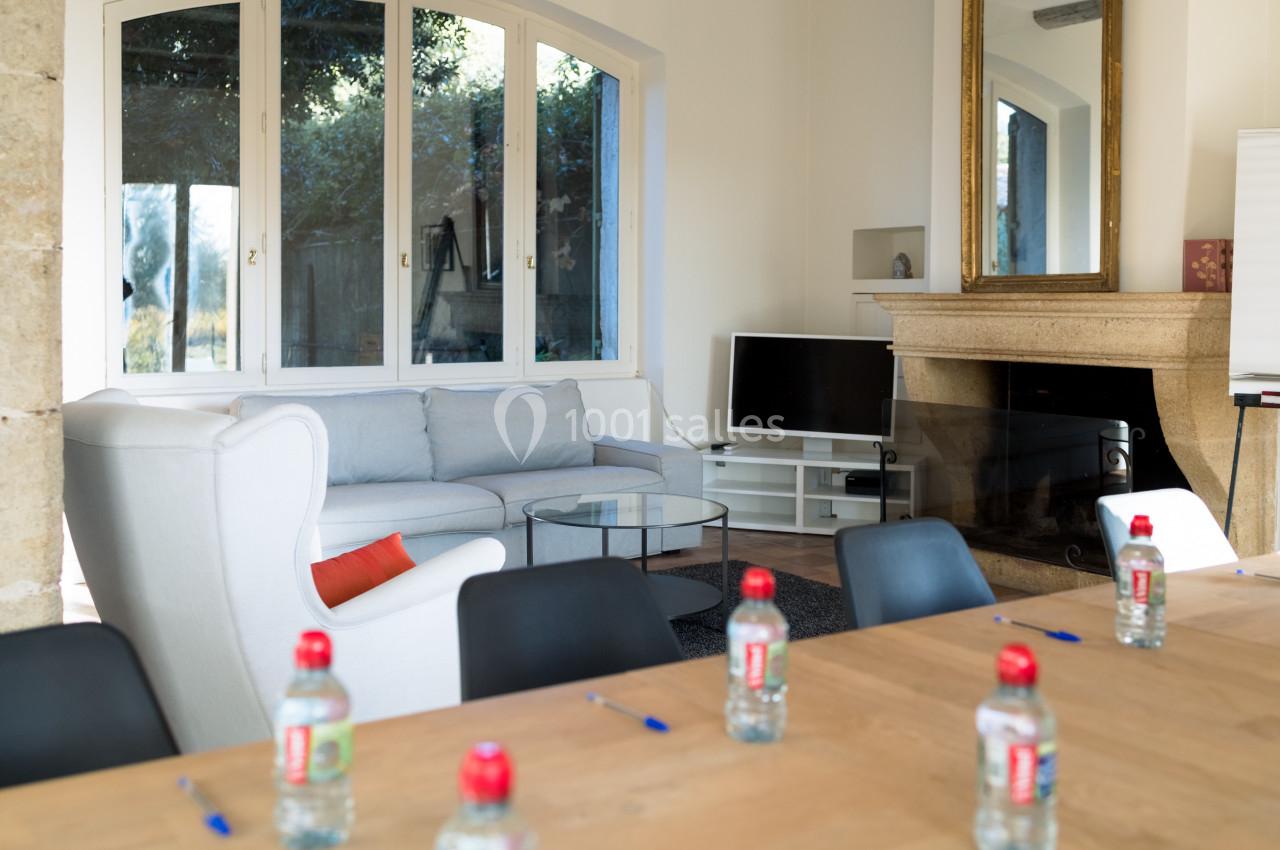 Salle lumineuse avec table en bois, chaises noires, canapé, cheminée, télévision et grandes fenêtres donnant sur un jardin.