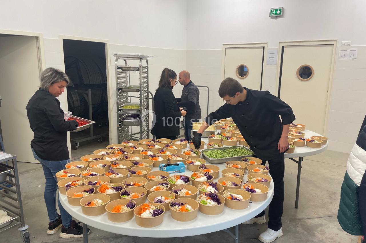 Des personnes préparent des salades colorées dans des bols en carton sur des tables dans une cuisine professionnelle.