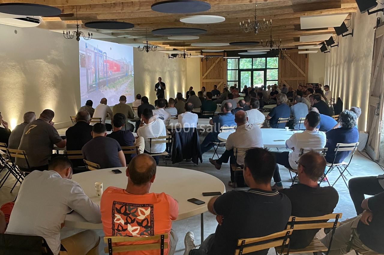 Des personnes assistent à une présentation projetée dans une salle avec un plafond en bois et des tables rondes.