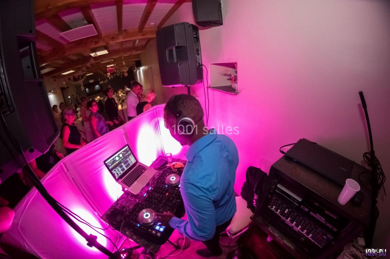 Un DJ mixe de la musique sur une console dans une salle éclairée par des lumières roses, avec des invités en arrière-plan.