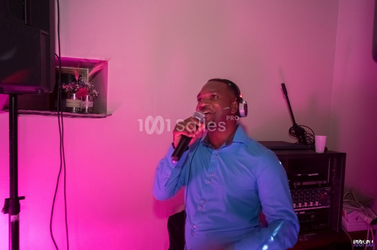 Un homme en chemise bleue chante au micro avec un casque audio, éclairé par une lumière rose dans une pièce.