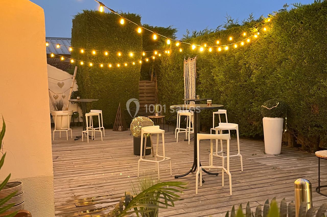 LA TERRASSE DU SALON GAUGUIN Terrasse en bois avec des guirlandes lumineuses suspendues, entourée de haies et aménagée avec tables et chaises blanches.