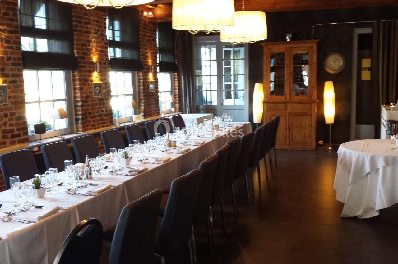 SALON MONET -40 PERSONNES Salle de réception avec longues tables dressées, vaisselle élégante et lumière naturelle entrant par des fenêtres.