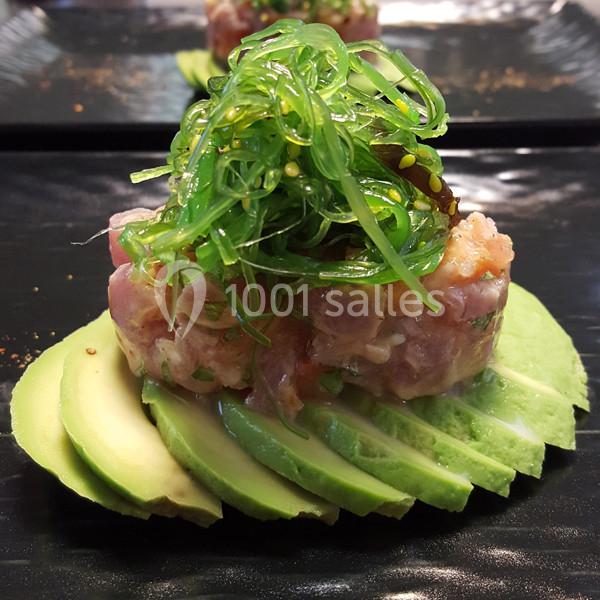 Tartare de poisson cru sur lit d'avocat, garni d'algues vertes, présenté sur une assiette noire.