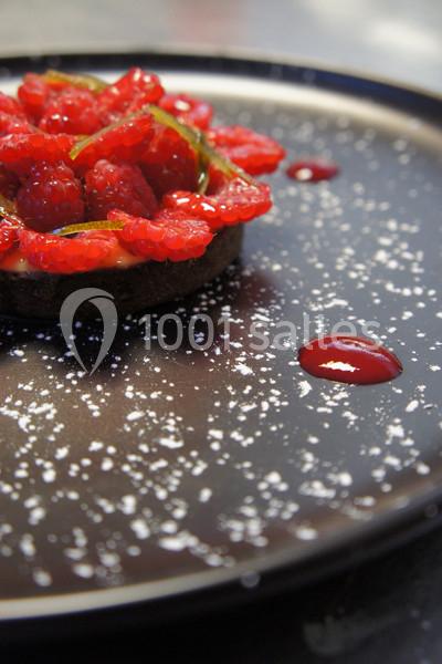 Tartelette garnie de framboises fraîches sur une assiette noire décorée de sucre glace et de gouttes de coulis.