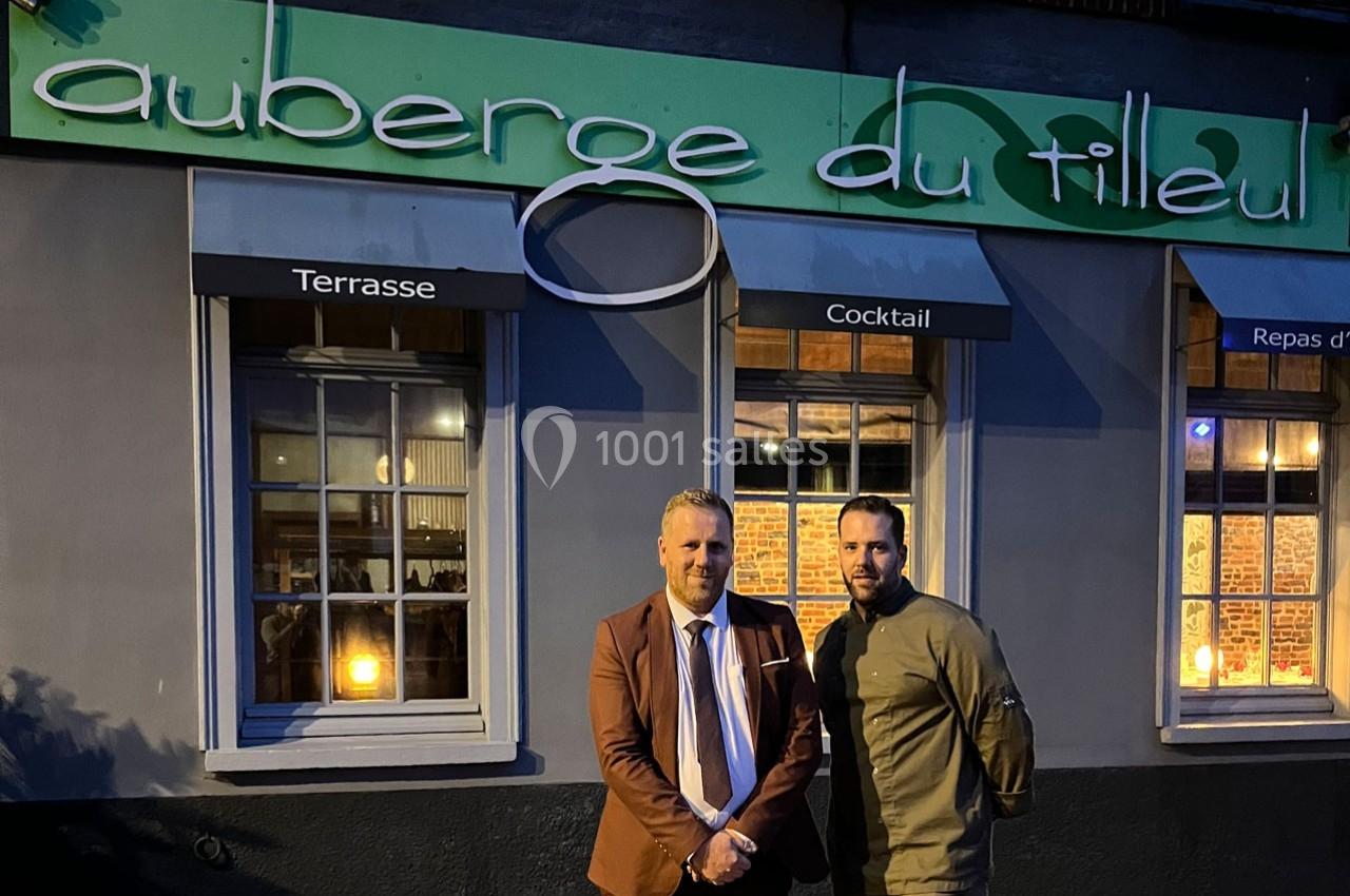Deux hommes posent devant la façade éclairée d'un restaurant nommé ’Auberge du Tilleul’ en soirée.