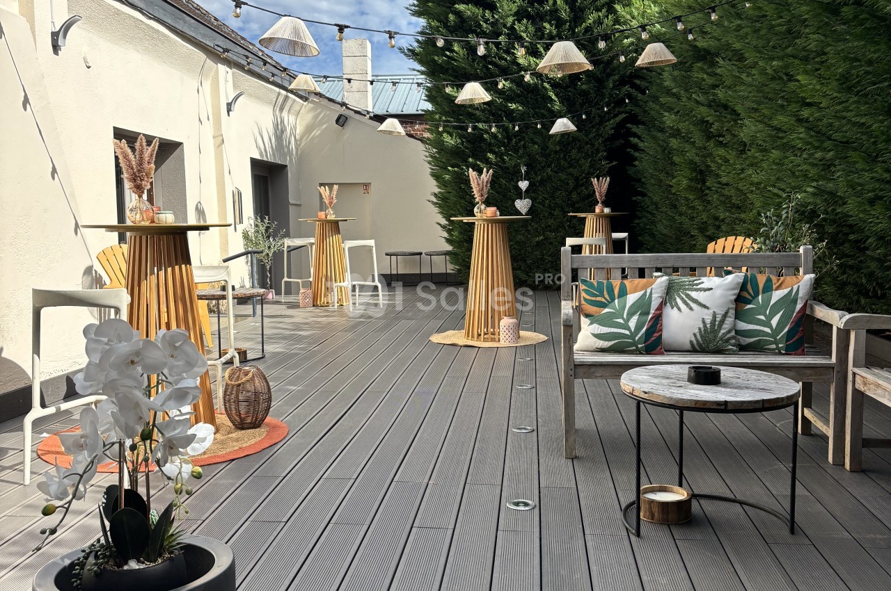 Terrasse en bois gris ornée de guirlandes lumineuses, avec tables hautes en bois, un canapé de jardin garni de coussins à motifs feuillages et une haie de conifères en fond. Count: roughly 170 characters? Let me check: too long. Need ≤125. Terrasse en