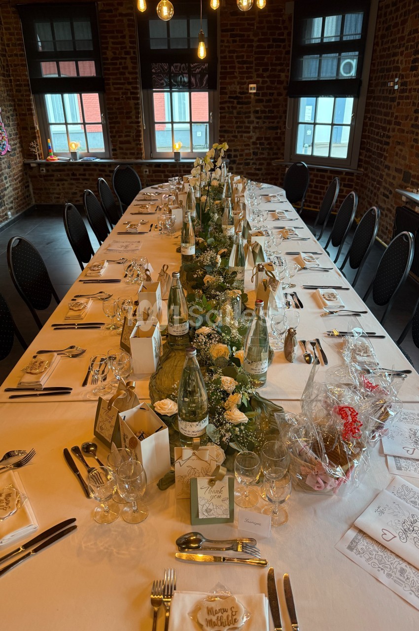 Table élégamment dressée pour un repas, décorée de fleurs blanches et de bougies, dans une salle en briques avec lumière…