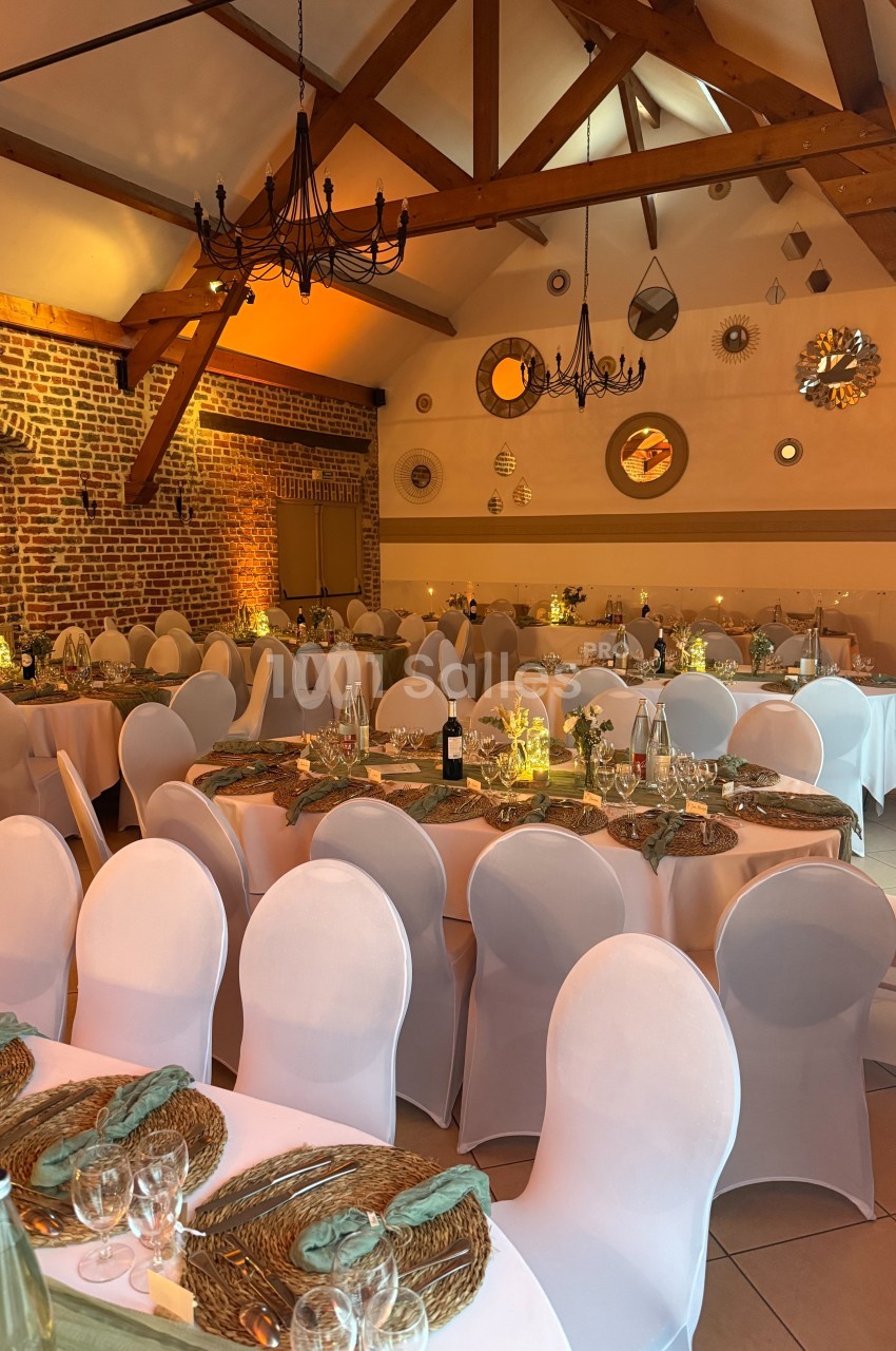 Salle de réception décorée avec des tables rondes, nappes blanches, chaises houssées et éclairage chaleureux.