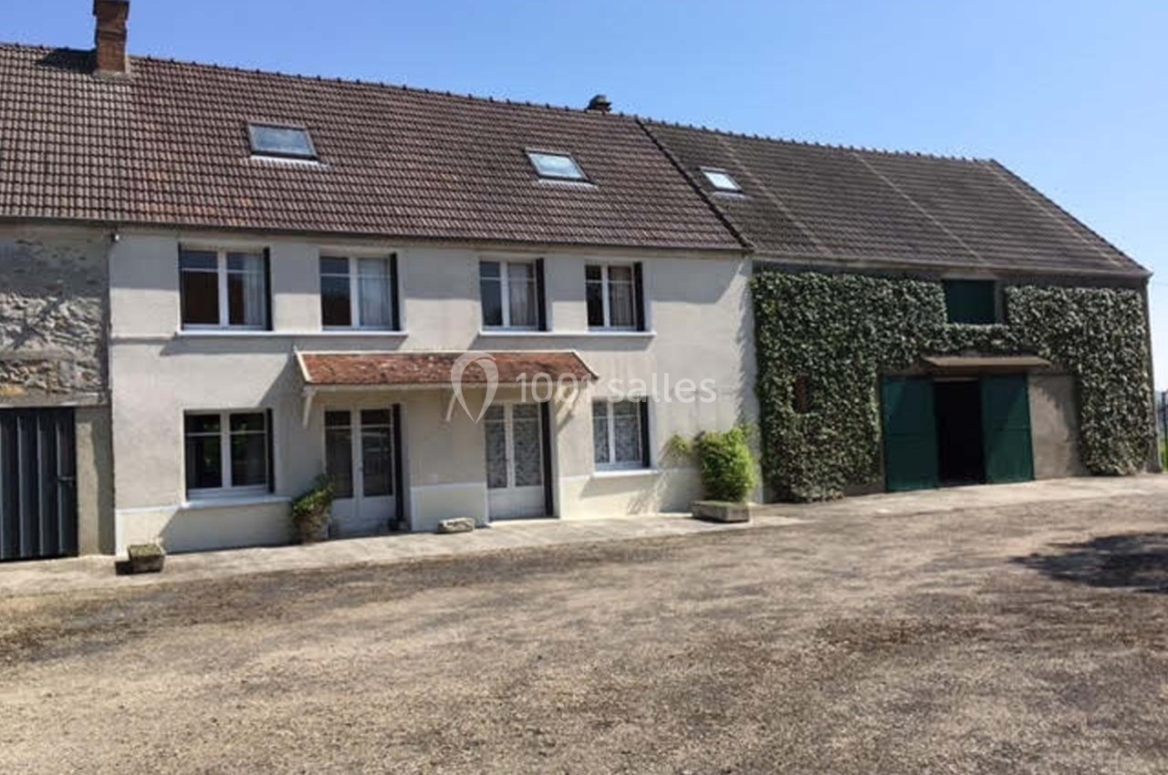 Maison de campagne avec façade claire, toit en tuiles et grange attenante recouverte partiellement de végétation.