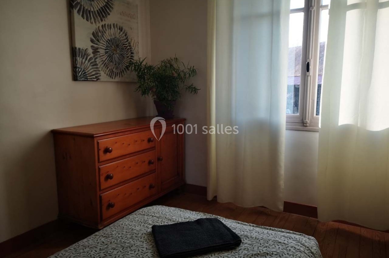 Chambre lumineuse avec une commode en bois, un lit recouvert d'un drap à motifs et une fenêtre avec des rideaux blancs.