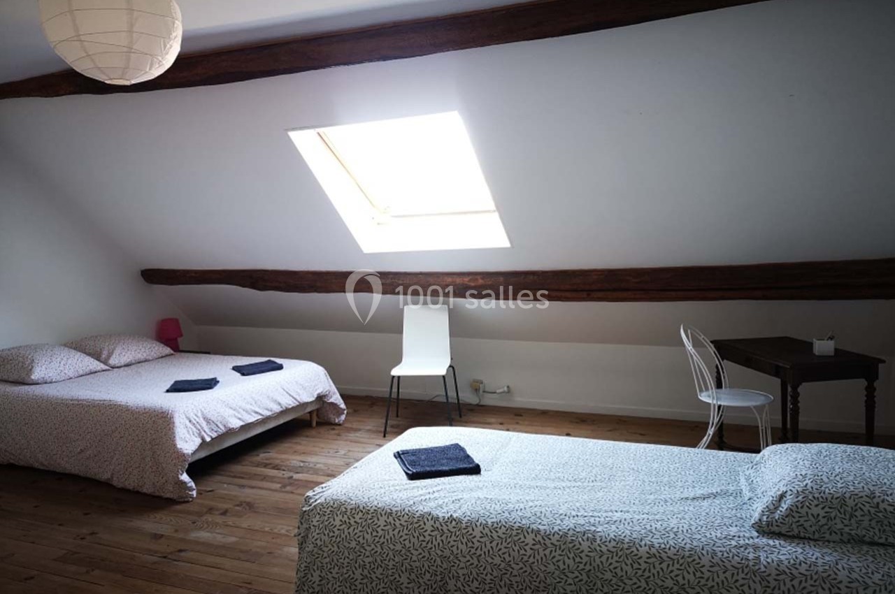 Chambre mansardée avec deux lits simples, un bureau en bois, une chaise blanche et une fenêtre de toit.