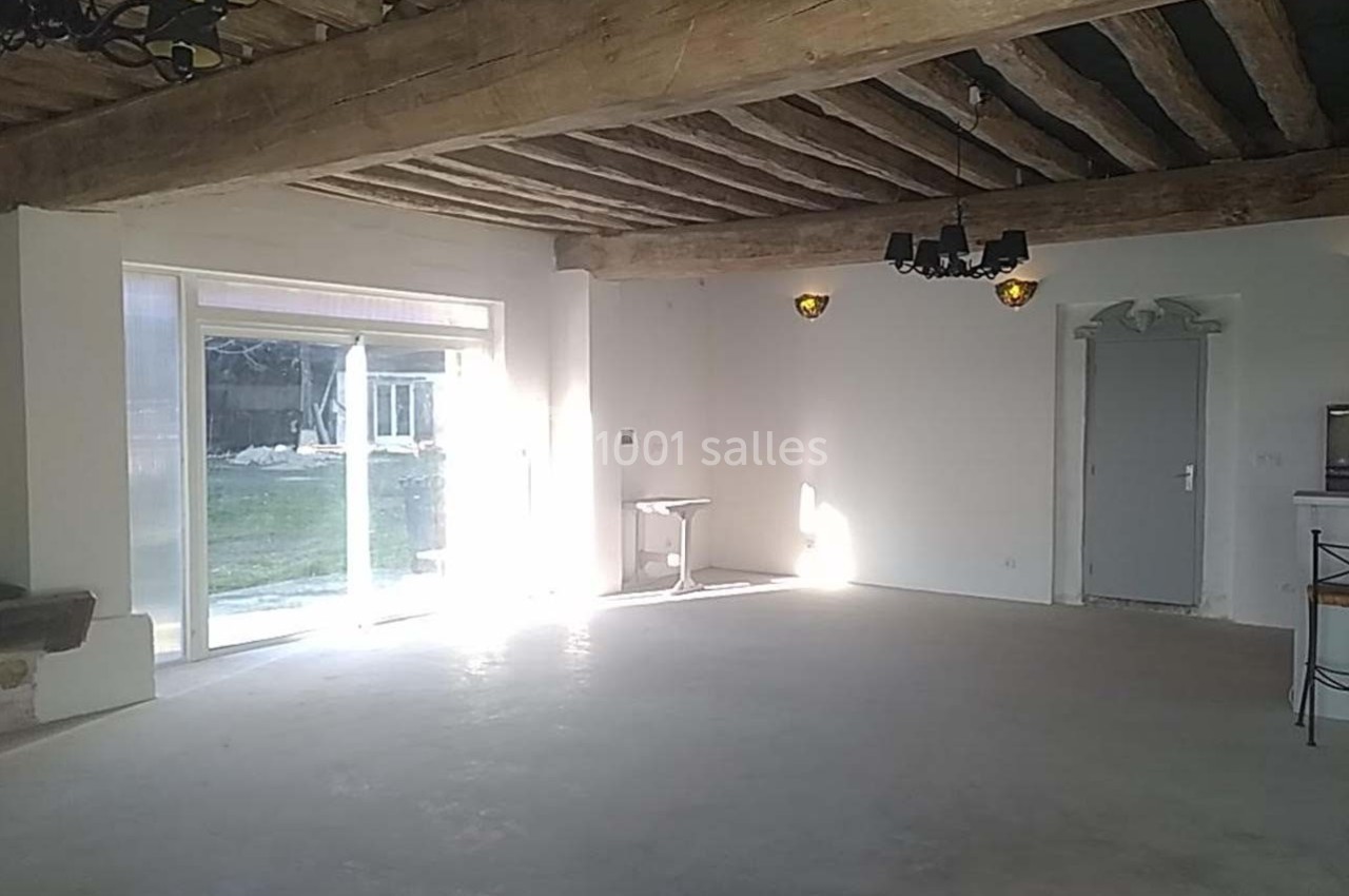 Grande pièce lumineuse avec poutres apparentes, sol en béton, porte et baie vitrée donnant sur un jardin.